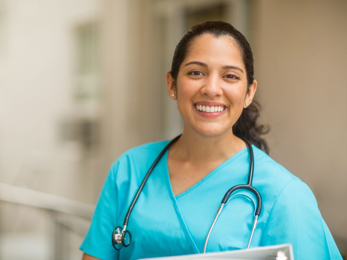 A female nurse smiles holding a clipboard and stethoscope.